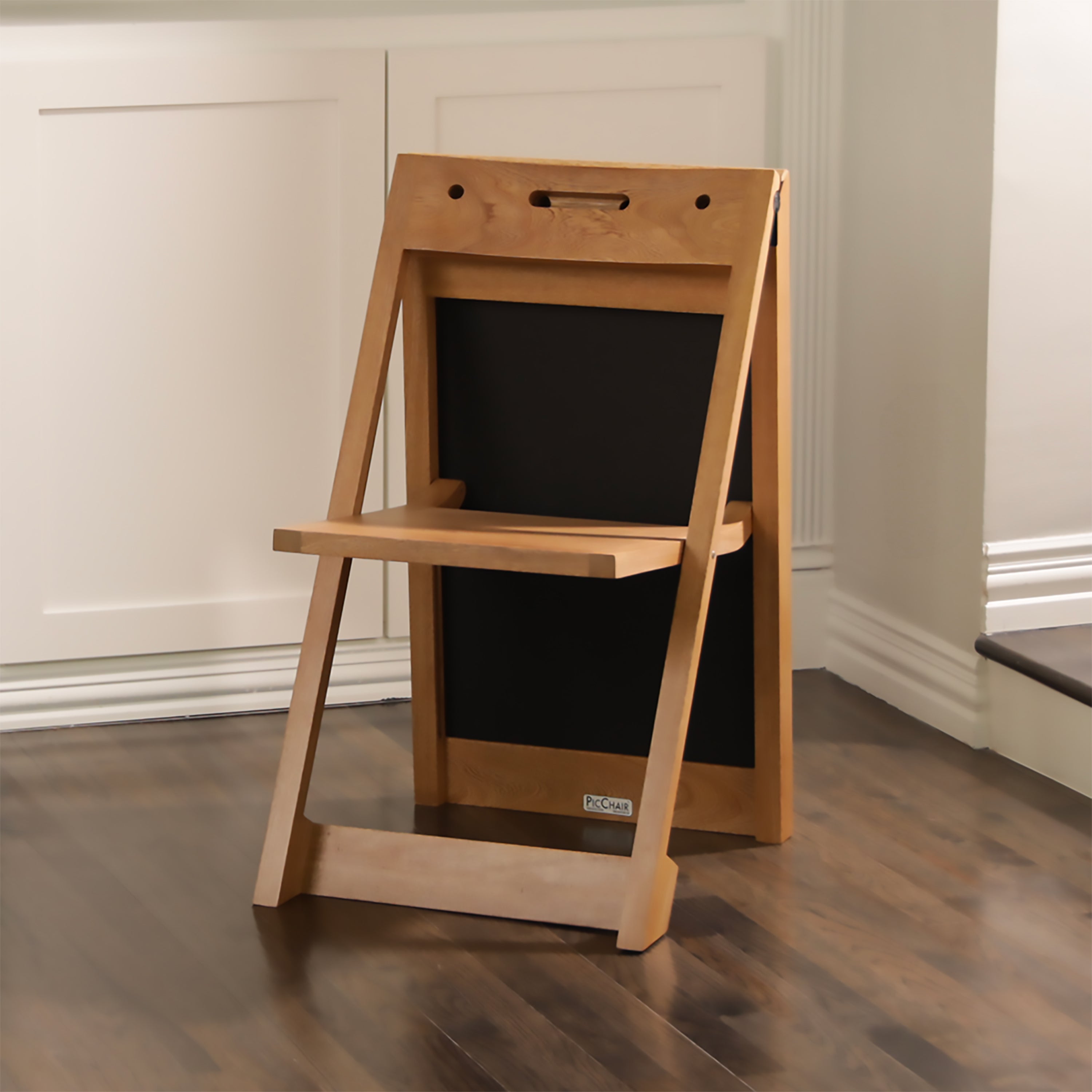 The PicChair in natural oak color on dark wooden floor with off-white paneled wall; front view;