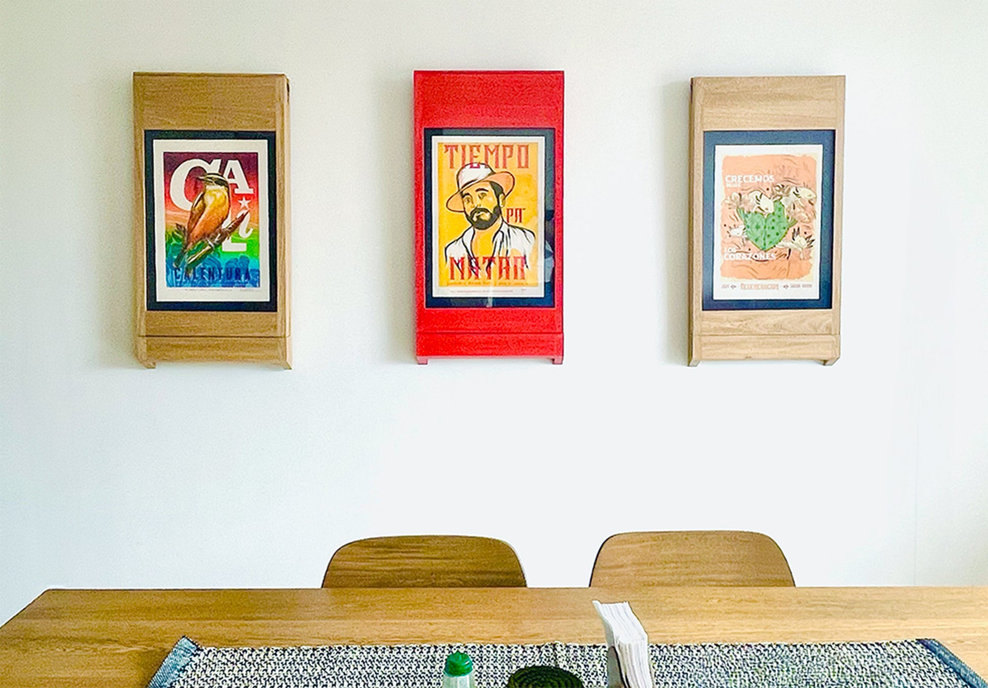 Three PicChairs hanging on off-white colored wall. each chair has a different art poster in it. There's also a wooden table in the foreground with two wooden chairs.