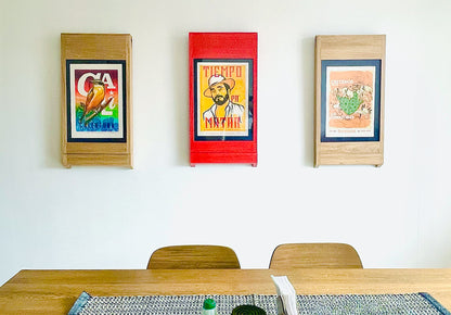 Three PicChairs hanging on off-white colored wall. each chair has a different art poster in it. There's also a wooden table in the foreground with two wooden chairs.