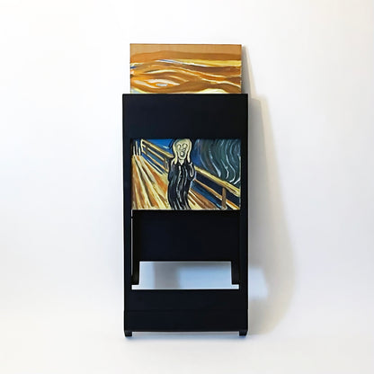 The PicChair in Black leaning against the wall with painting of The Scream halfway  in the picture frame.