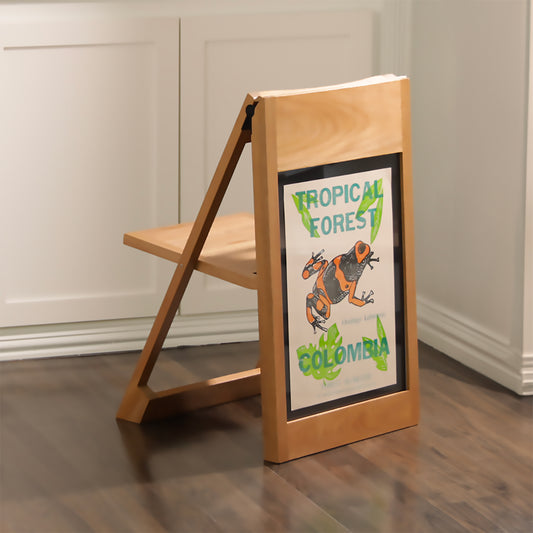 The PicChair in natural oak color on dark wooden floor with off-white paneled wall; Poster of a graphic illustration of a frog with words "Tropical Forest; Colombia".